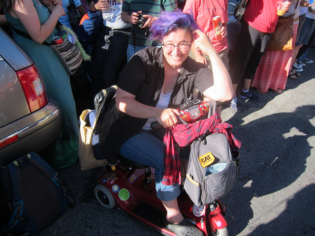 A purple-haired white woman smiling while sitting on her mobility scooter in front of a small crowd of women