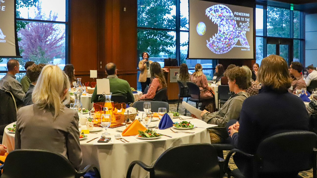 Ruth Jebe introduces the Fall 2023 Business Ethics Roundtable on "PIONEERING SOLUTIONS TO THE PLASTIC CHALLENGE," with attendees seated at tables focused on the presentation. The room is bathed in natural light, emphasizing the professional and attentive atmosphere of the event.