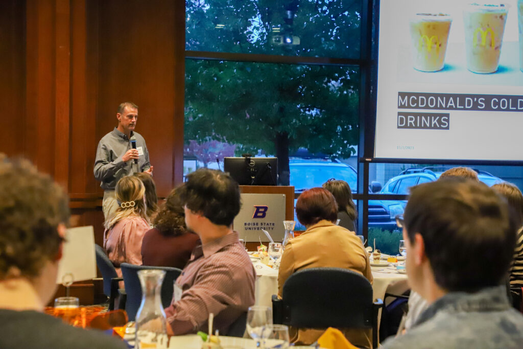 Darren Kyle presents on the challenges of plastic disposal at his McDonald's franchises at the Fall 2023 Business Ethics Roundtable at Boise State University, addressing an audience seated at tables with orange cloths in a room with a view of trees through large windows.