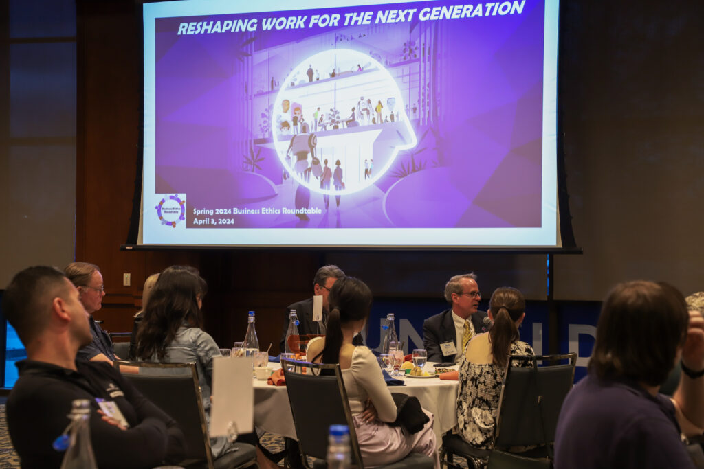This image shows a conference room where participants are seated at round tables, engaged in discussion. At the front, there is a large presentation screen displaying the title "RESHAPING WORK FOR THE NEXT GENERATION", with the subheading "Spring 2024 Business Ethics Roundtable, April 3, 2024". The screen also features a graphic of diverse individuals in a workplace setting. In the foreground, some participants are facing away from the camera, drawing attention to the conversational and interactive nature of the event. The environment suggests a professional gathering focused on discussing the future of business ethics and work.