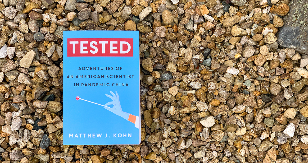 A book sitting on some rocks with the title, "Tested: Adventures of an American Scientist in Pandemic China" by Matthew J. Kohn