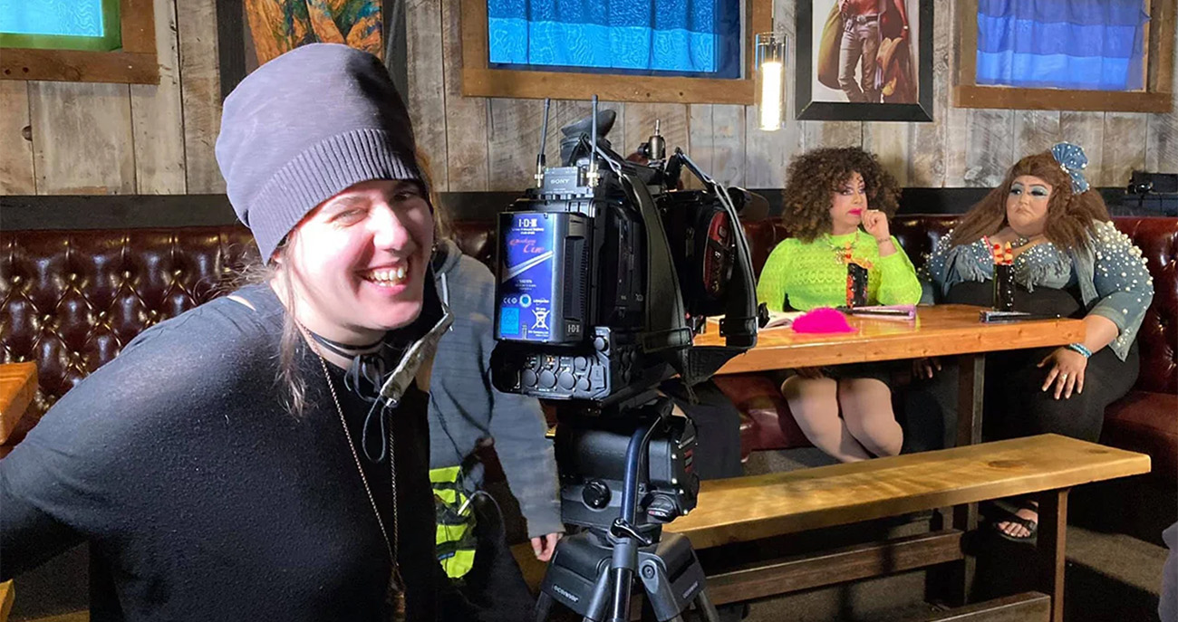 A film student directs two actors on set in a bar with a cinema camera next to her