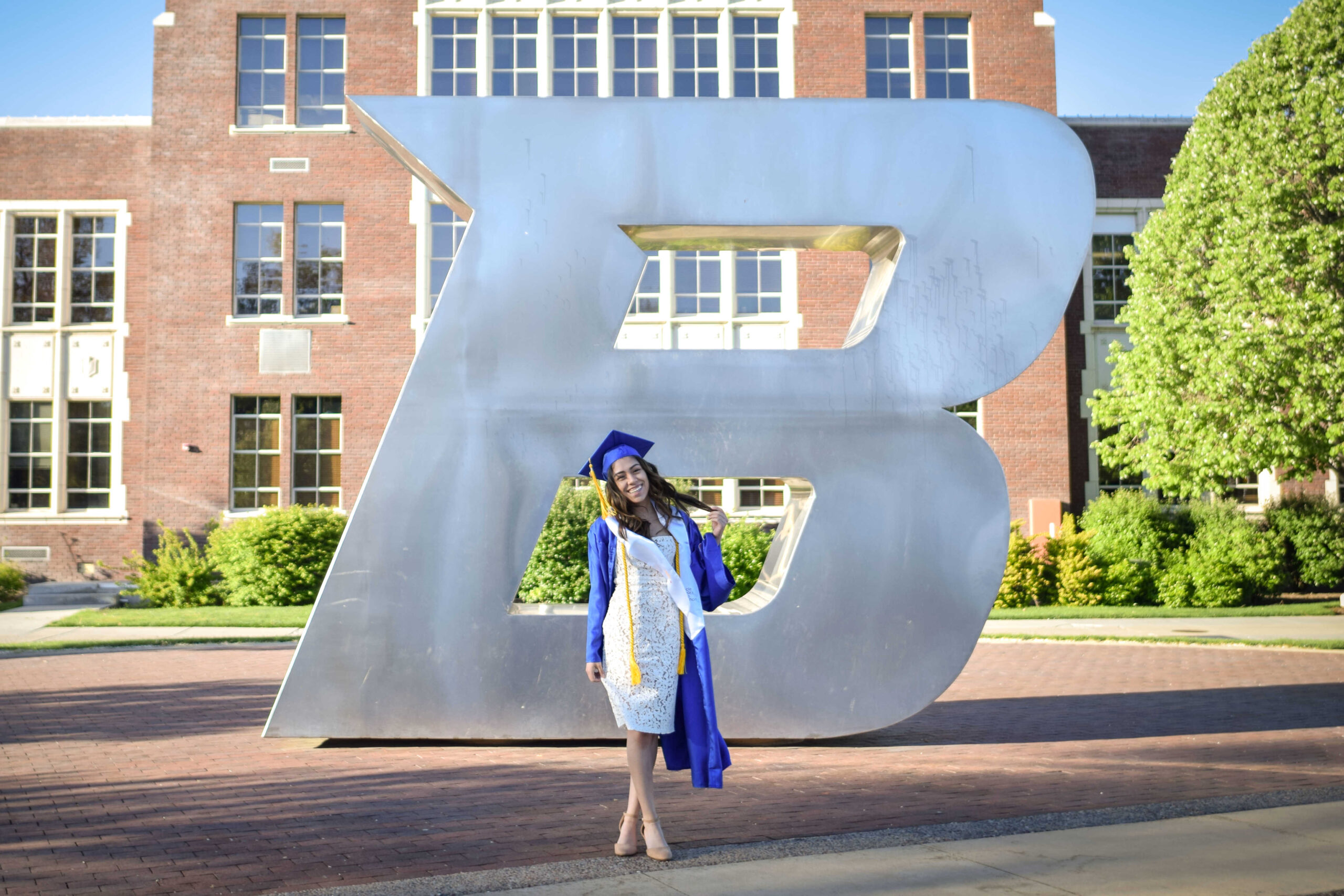 Graduating student posing in front of the Boise State B, wearing a white dress, open blue graduation gown, and graduation cap