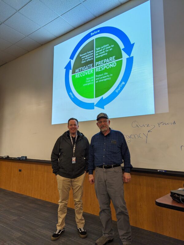 Photo of Crash Marusich, the Deputy Director for Ada County Office of Emergency Management and Community Resilience, and Ben Wells, Boise State's emergency manager, standing side by side in front of the projector image.