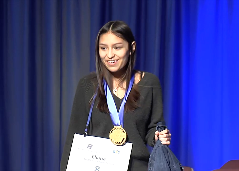 young woman smiles and stands on stage with medal around her neck and certificate in hand