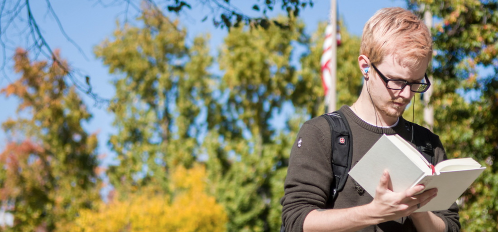 A student reading a book with trees and an american flag in the background