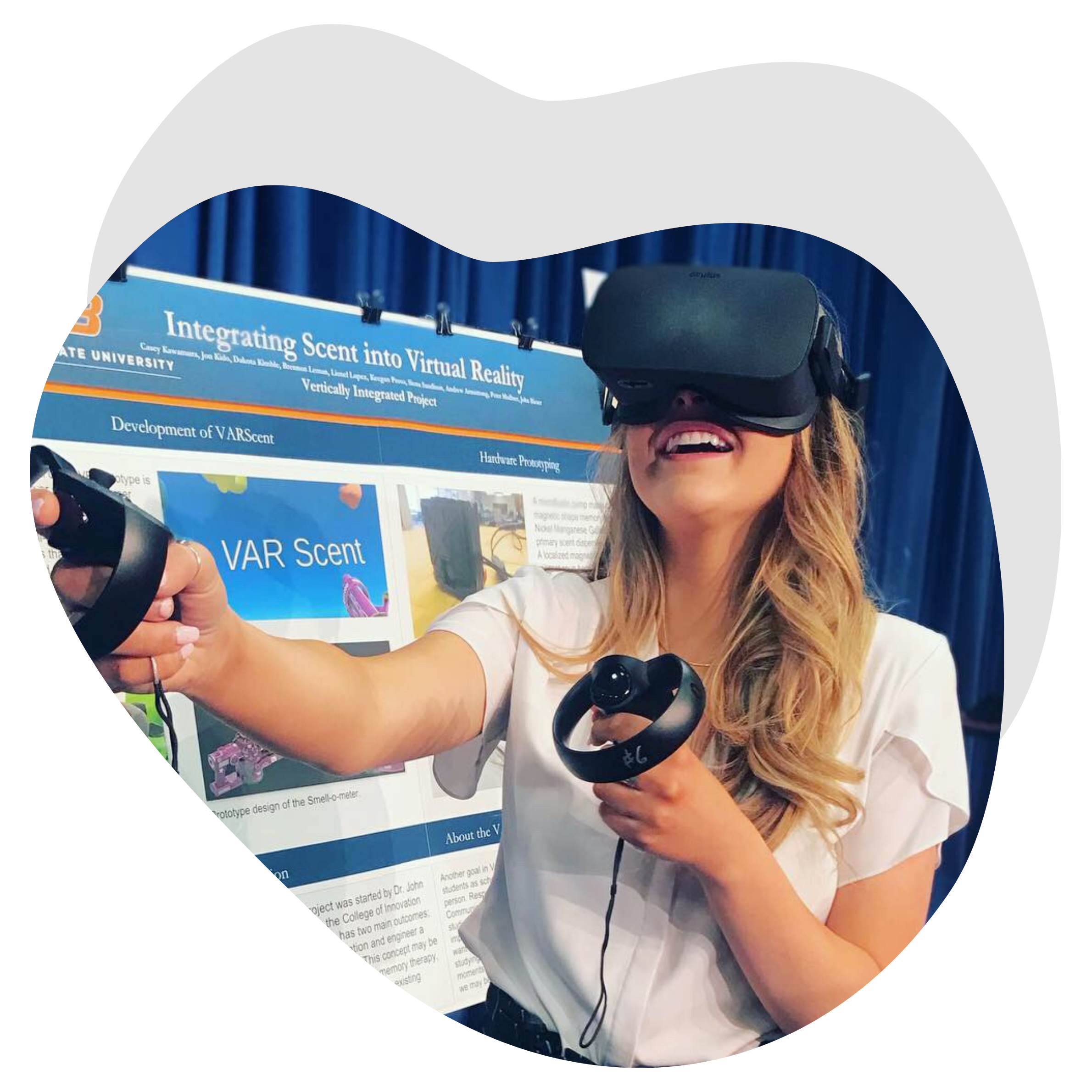 photo of an undergraduate student demonstrating her research using a virtual reality headset. The student stands in front of a research poster at a poster session. The photo is cropped into an organic blob shape with a gray drop shadow