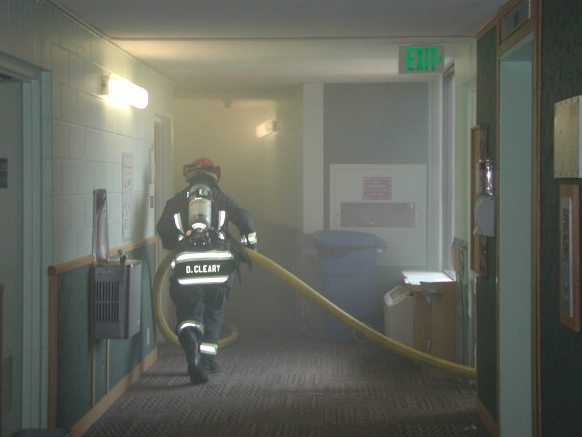 Firefighter carrying a large hose down a smokey hallway.