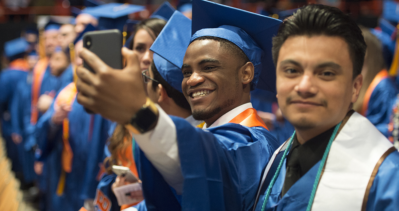 line of Boise State graduates in cap and gowns with one graduate taking a selfie