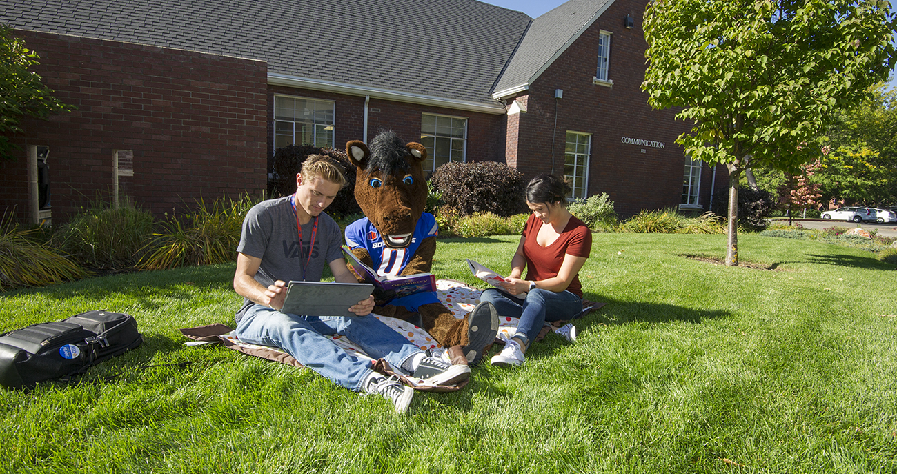 two Boise State students and Buster Bronco sitting on the grass in front of a building on campus