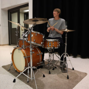 Student smiles will sitting on a stool, casually holding drumsticks behind a drumset. 