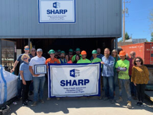 Employees of Nucor Harris Rebar stand outside their facility with a Safety and Health Achievement Recognition Program flag that recognizes their exemplary safety and health achievements. Also pictured are team members from the Idaho Occupational Safety and Health Consultation Program.