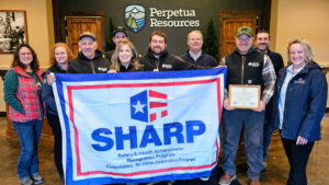 Employees of Perpetua Resources stand in the lobby of their facility with a Safety and Health Achievement Recognition Program flag that recognizes their exemplary safety and health achievements. Also pictured is the Idaho Occupational Safety and Health Consultation Program's Manager.