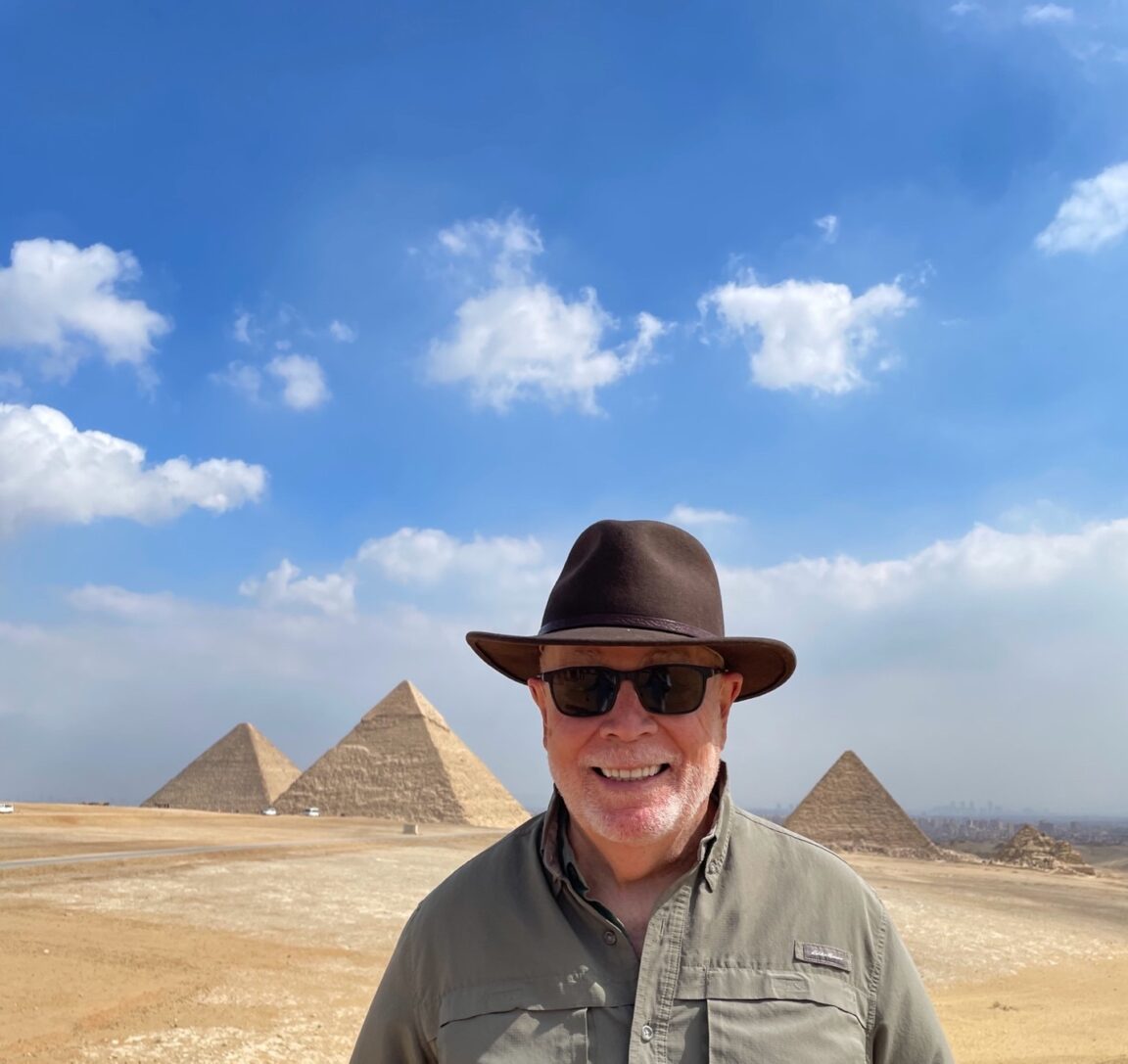 Tom wearing a hat and sunglasses stands in front of pyramids with a blue sky and white clouds in the background