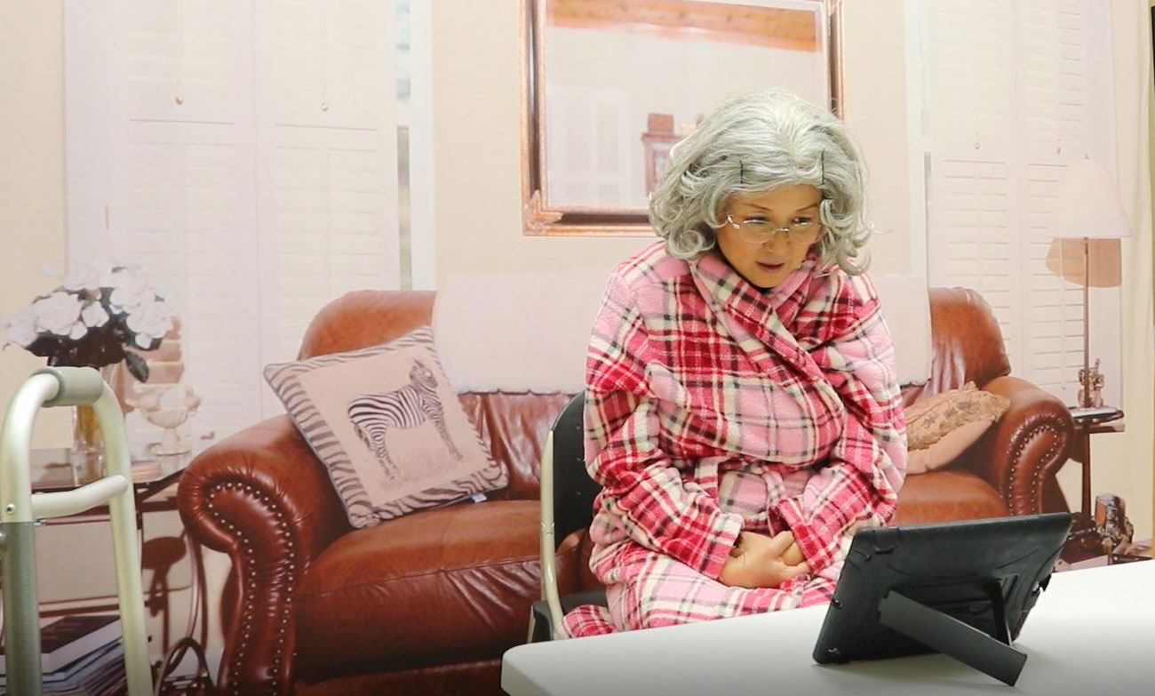 Janet Lo wears a grey wig and pink bathrobe, sitting at a table talking to a propped-up electronic tablet.