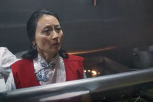 Janet Lo wears a red vest looking shocked in a dimly lit kitchen.
