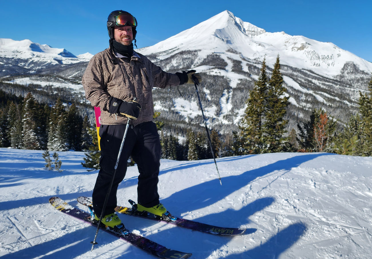 Justin Malinosky stands in front of a mountain smiling and wearing skis.