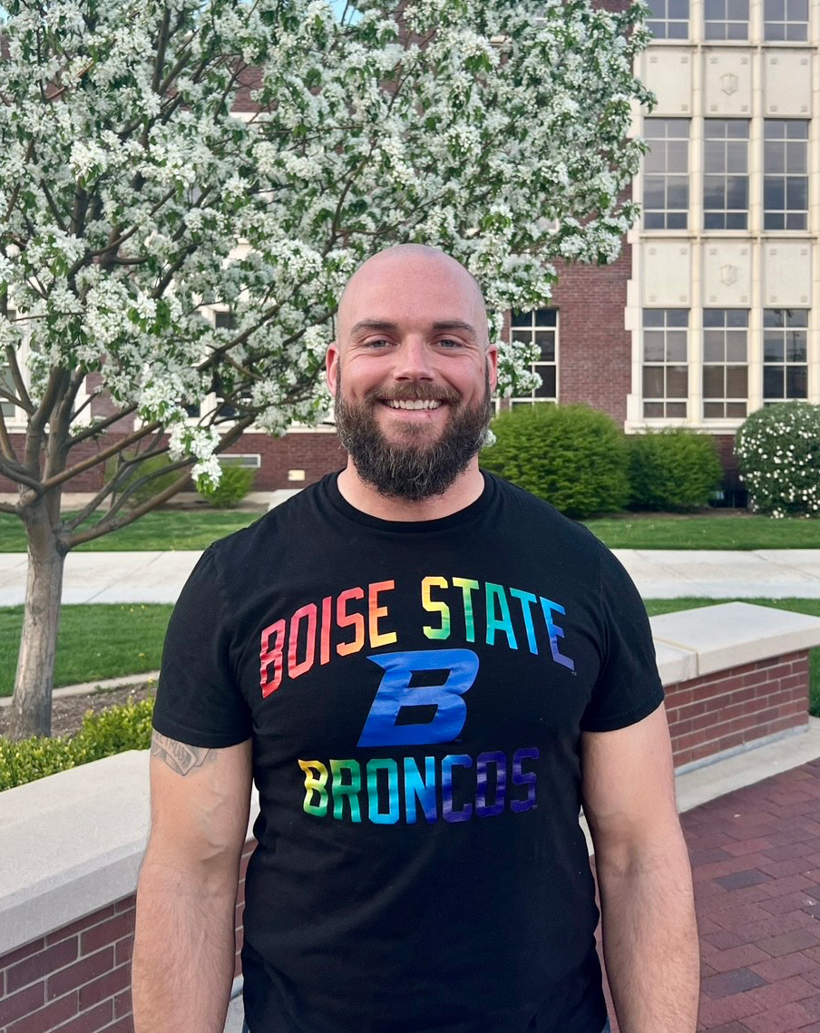 Chris Hughes wears a Boise State University tee shirt and smiles by a blooming tree.