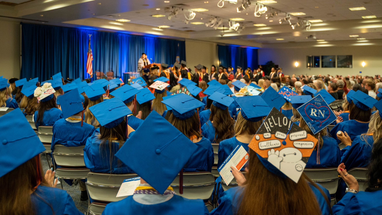 Blue caps and gowns fill the foreground as students sit in rows in front of the stage during Convocation.