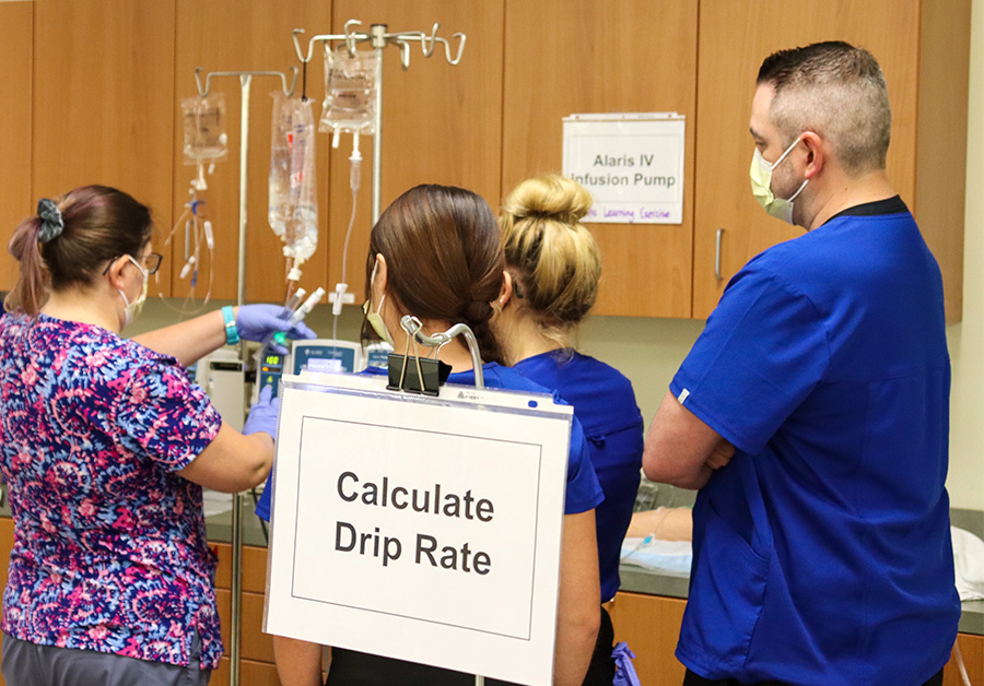 A professor demonstrates how to calculate drip rate in an IV skills lab to three students.