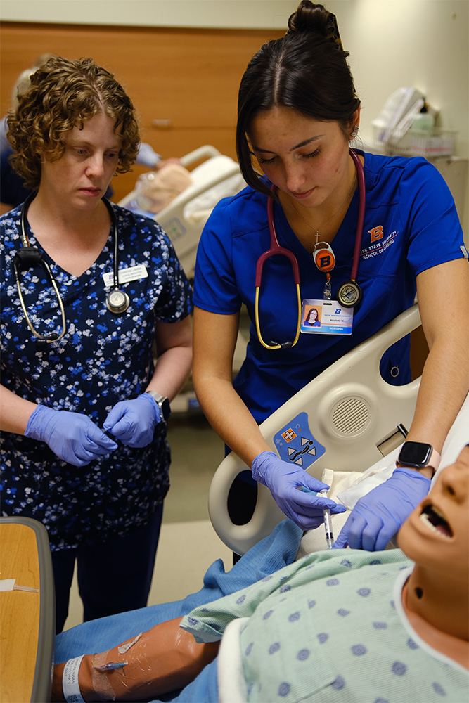 A professor looks on as a student practices an injection on a manikin.