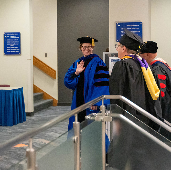 Kelley Connor wears her doctoral regalia to attend Convocation and waves at the camera from the top of a staircase.