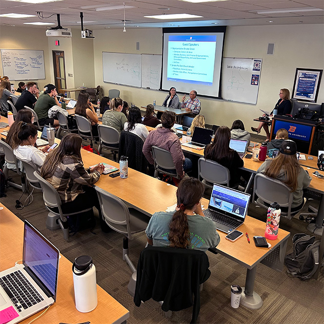 Students fill a lecture hall and listen to two guest speakers by a powerpoint presentation at the front of the room.