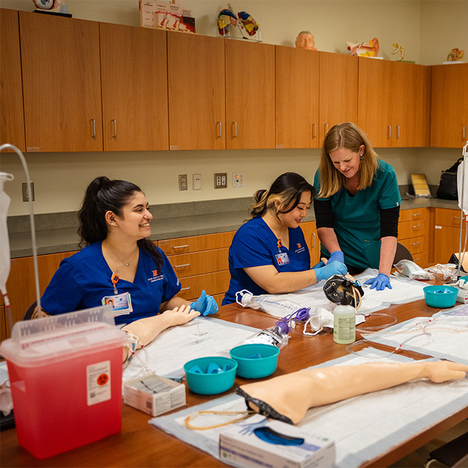 Two nursing students practice placing IVs in manikin arms while a nurse faculty assists.