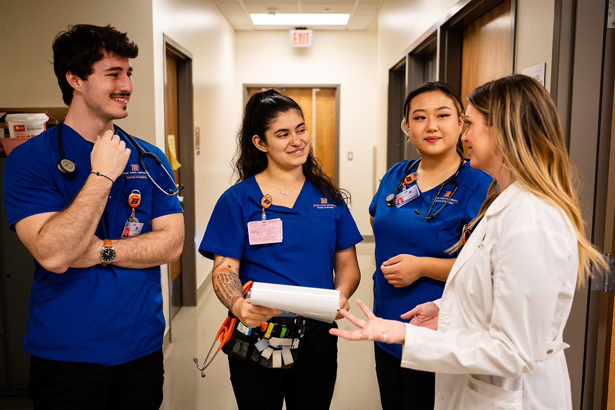 Three nursing students wearing blue scrubs stand in a hallway and conference with a nurse in a white coat.