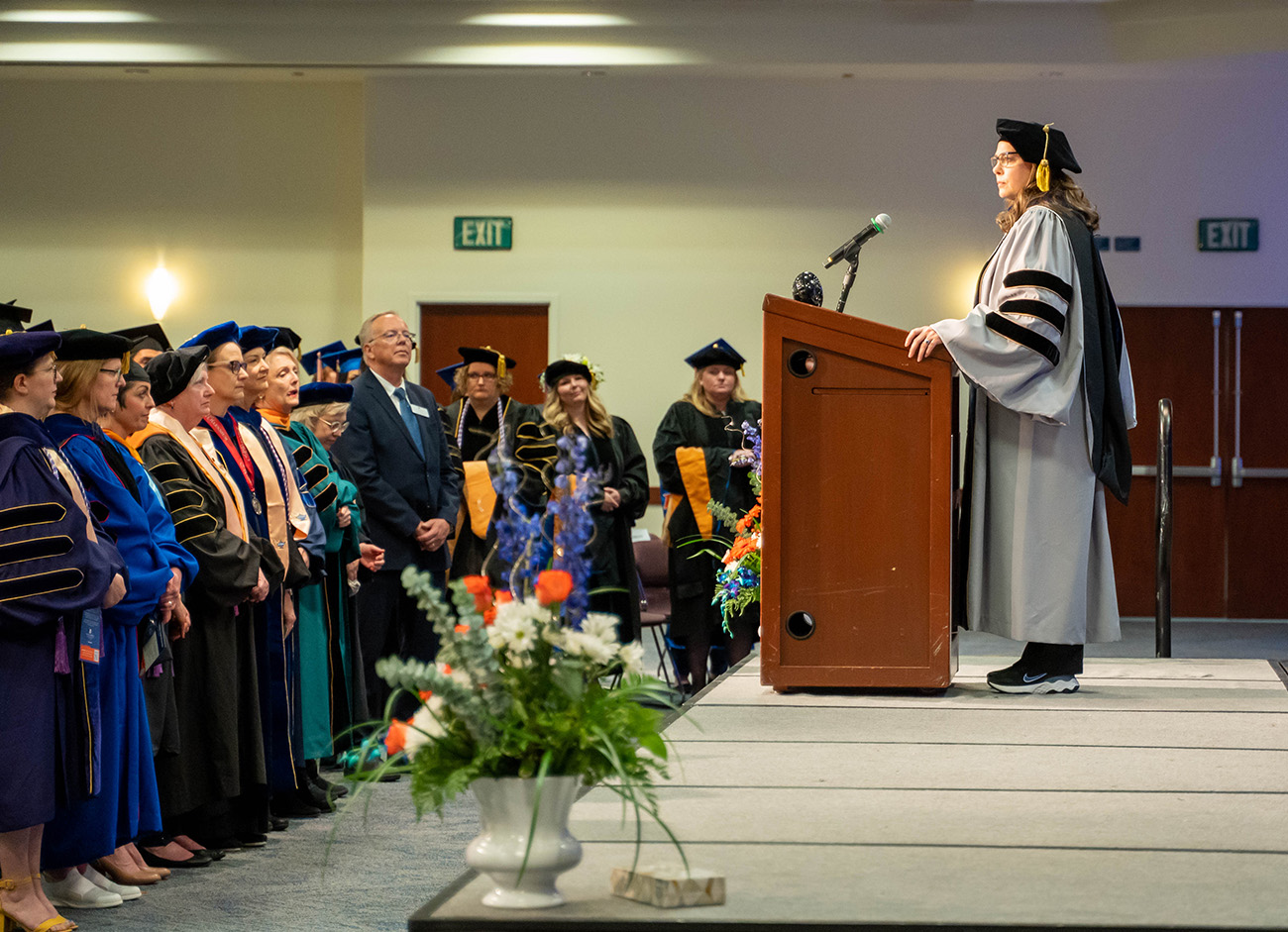 Poole stands at a podium in her doctoral regalia and addresses the faculty in their regalia standing in front of her.