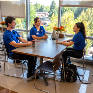 Three nurses talk and laugh together while sitting at a table by a wall of windows.