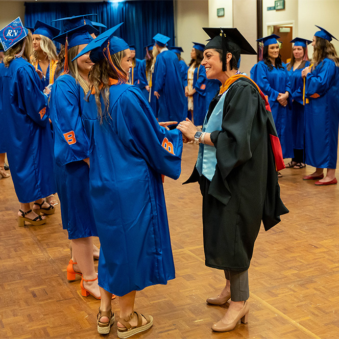 Angie wears her regalia and smiles as she shakes the hand of one student in a line of students all wearing their blue regalia.