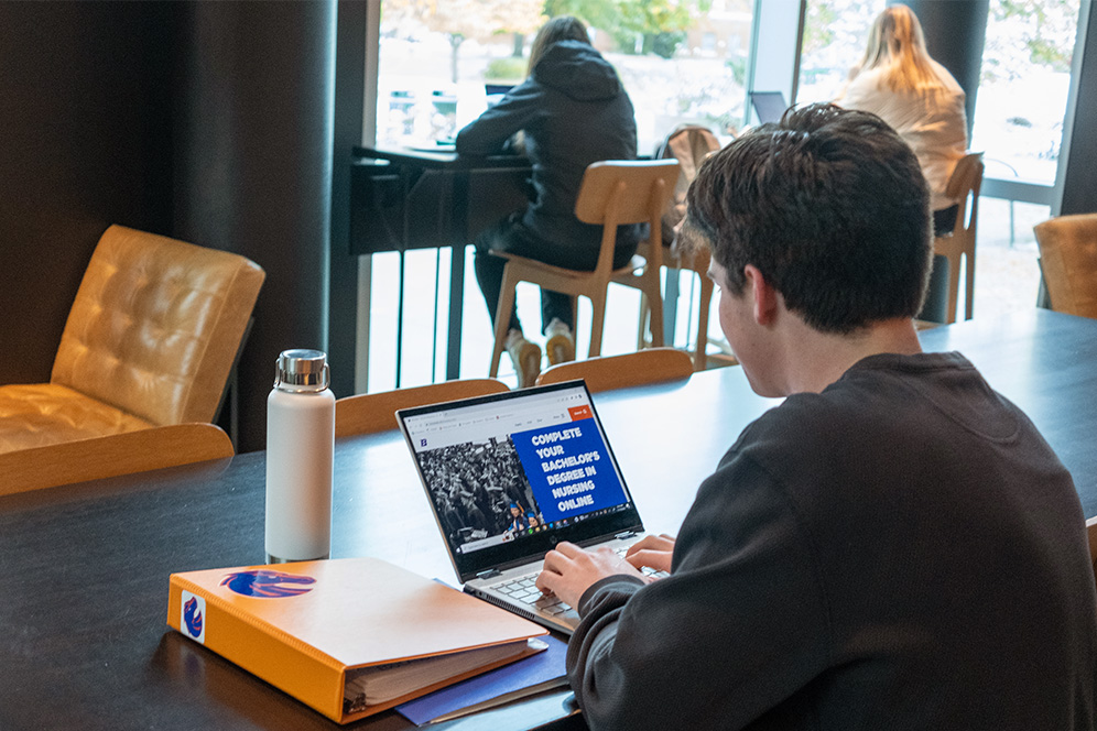 A student faces away from us while sitting in a coffee shop working on a laptop that reads "Complete you bachelor's degree in nursing online".