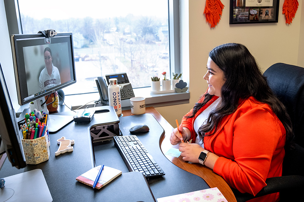 An advisor in an orange blazer sits at her desk and talks to a student through a video call on her computer.