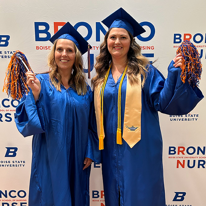 Two students wear blue and orange regalia and hold pompoms in front of a Bronco Nurse backdrop. Their stoles are apricot and have a lamp stitched with the word "Nursing".