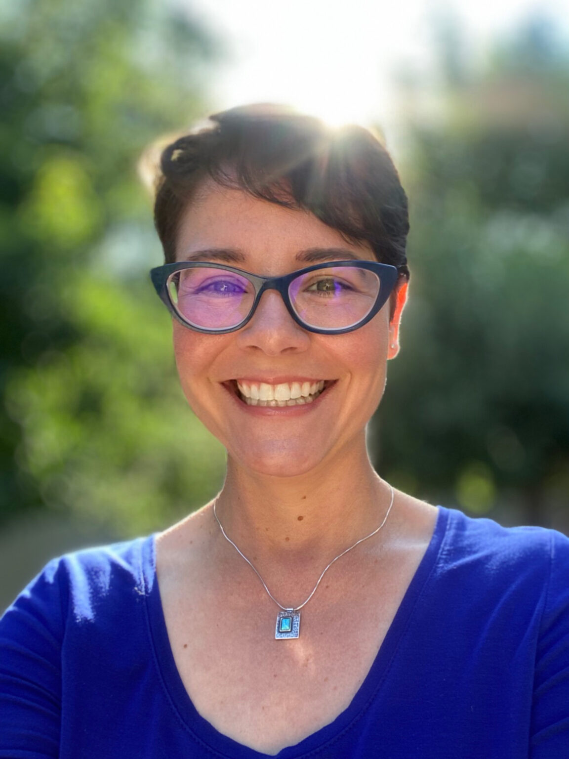 Picture of Ti Macklin smiling. Ti is a white woman with cropped brown hair. She is wearing blue framed glasses, a blue shirt, and is standing in front of a blurred background of nature.